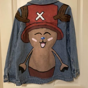 May include: A denim jacket featuring a hand-painted cartoon character design. The design includes a red hat with a white cross, brown antlers, and a smiling face with pink cheeks. The jacket is hanging on a white hanger.