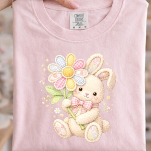 May include: A light pink t-shirt featuring a watercolor illustration of a bunny holding a flower. The bunny has a pink bow and the flower has pastel gingham petals. The text "INCLUDES: PNG FILE" is at the bottom.