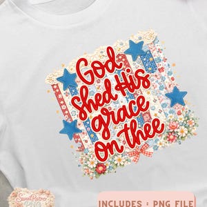May include: White t-shirt featuring a patriotic design with the text "God shed His grace on thee" in red script. The design includes blue stars, floral accents, and a patchwork pattern.