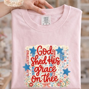 May include: A light pink t-shirt with the phrase "God Shed His grace On thee" in red script. The design includes blue stars, floral accents, and a patriotic theme. The shirt is folded and held by a hand.