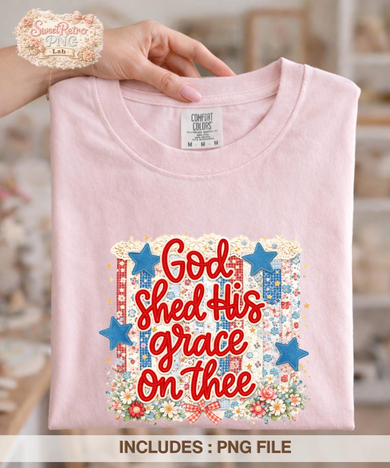 May include: A light pink t-shirt with the phrase "God Shed His grace On thee" in red script. The design includes blue stars, floral accents, and a patriotic theme. The shirt is folded and held by a hand.