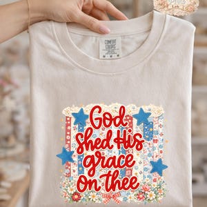 May include: A beige Comfort Colors t-shirt featuring the text "God Shed His grace On thee" in red script. The design includes blue stars, floral accents, and a patriotic color scheme.