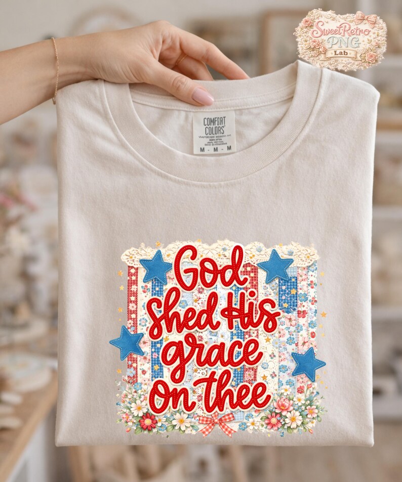 May include: A beige Comfort Colors t-shirt featuring the text "God Shed His grace On thee" in red script. The design includes blue stars, floral accents, and a patriotic color scheme.