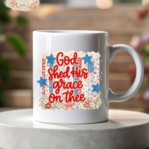 May include: White ceramic mug with a patriotic design. The mug features the text "God shed His grace on thee" in red script, surrounded by blue stars, floral patterns, and a red, white, and blue color scheme.