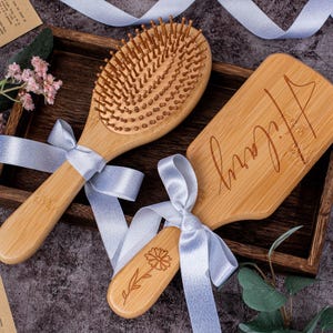 May include: Three wooden hairbrushes with light blue ribbon bows. One brush is oval with bristles, another is rectangular with the name "Hilary" engraved, and the third is small with a flower design. They are arranged in a wooden tray.