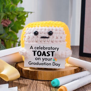 May include: A crocheted figure with yellow hair and a white face holds a sign that reads "A celebratory TOAST on your Graduation Day." The figure is on a wooden slice, with markers and decorative elements in the background.