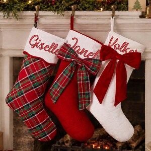 Peut inclure: Trois chaussettes de Noël suspendues en rangée. La première est à carreaux rouges et verts avec un revers blanc brodé du nom Giselle. La deuxième est rouge uni avec un nœud à carreaux et le nom Janet. La troisième est blanche avec un nœud rouge et le nom Volker.