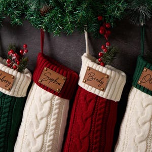 May include: Four cable-knit Christmas stockings in alternating red, green, and white colors. Each stocking features a rectangular brown leather patch with a name engraved in black script: Hubery, Sophia, Brady, and Olivia, with a paw print icon.