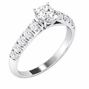 Könnte beinhalten: Ein silberner Verlobungsring mit einem großen, rund geschliffenen Diamanten im Zentrum. Das Band ist mit kleineren Diamanten besetzt, die für zusätzlichen Glanz sorgen. Der Ring ist in einer klassischen Krappenfassung gehalten, die die Brillanz der Steine hervorhebt.