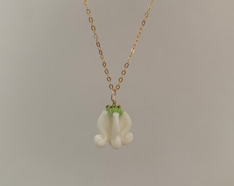 Puede incluir: Un delicado collar de cadena dorada con un colgante de flor de porcelana blanca. La flor tiene un centro verde y es una representación realista de una flor.