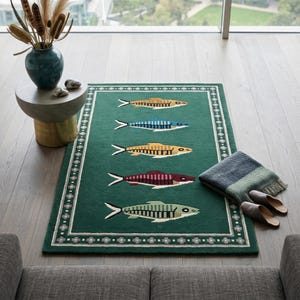 Puede incluir: Una alfombra rectangular verde con un borde blanco y gris con un patrón floral repetido. La alfombra muestra cinco diseños de peces coloridos. Una manta azul y verde y un par de zapatillas grises están al lado de la alfombra.