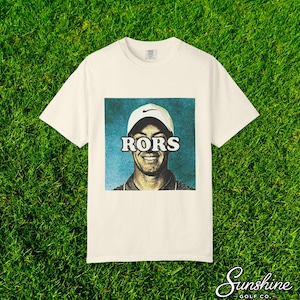May include: Cream-colored t-shirt featuring a graphic of a smiling person wearing a white cap with the Nike logo. The word "RORS" is printed across the person's eyes. The shirt is on a green grass background, with the Sunshine Golf Co. logo.