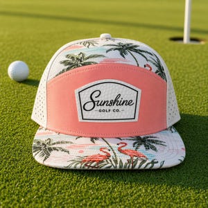May include: A white and pink golf hat with a tropical design featuring palm trees and flamingos. The hat has a pink front panel with the text "Sunshine - Golf Co." and a white mesh back. A golf ball is visible.