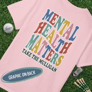May include: A light pink t-shirt with the words "MENTAL HEALTH MATTERS" in colorful, retro-style lettering. Below, it says "TAKE THE MULLIGAN". The shirt has a graphic on the back.