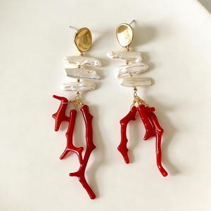 Puede incluir: Pendientes colgantes con tachuelas doradas, perlas blancas irregulares y abalorios rojos con forma de coral. Los pendientes se muestran sobre una superficie blanca, mostrando su diseño elegante.