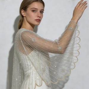 以下が含まれることがあります： 繊細なシルバービーズと輝く装飾が施された、アイボリーのシアーケープ。ケープは、金色のトリムが付いたスカラップエッジを備えています。デザインはエレガントで流れるようで、ブライダルやフォーマルな機会に適しています。美しいアクセサリーです。