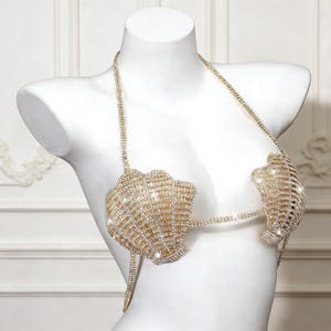 May include: A gold-colored rhinestone bikini top displayed on a white mannequin. The top features a seashell design with sparkling rhinestones and delicate chain straps. The bra is designed to be worn as a top.