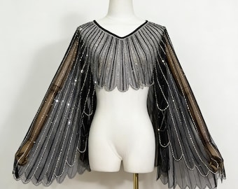 Può includere: Mantella nera e argento con scollo a V. La mantella presenta un orlo smerlato ed è adornata da linee verticali di paillettes argentate. Le maniche trasparenti si estendono dalle spalle, creando un look drammatico ed elegante.