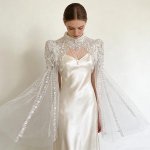 Op de afbeelding: Een witte satijnen trouwjurk met een hooggesloten, geborduurde cape. De cape heeft ingewikkeld kralenwerk en loopt uit in transparante, vloeiende mouwen. Een delicate ketting maakt de jurk compleet. Het ontwerp is elegant.