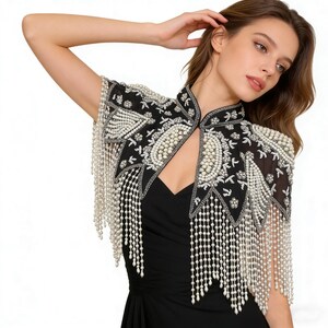 Op de afbeelding: Een zwarte geborduurde bolero met witte parelaccenten en franjes. De bolero heeft ingewikkelde bloemen- en geometrische patronen. Het kledingstuk wordt over een zwarte jurk gedragen, wat een elegante, formele look creëert.