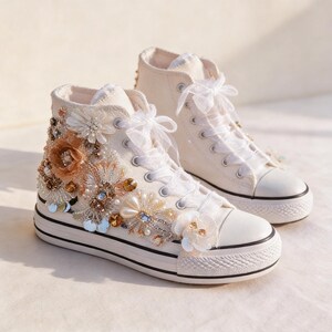 May include: A pair of ivory high-top sneakers adorned with floral embellishments. The sneakers feature white laces, a black stripe on the sole, and decorative beading and sequins. The floral accents include pearls and gold-toned accents.