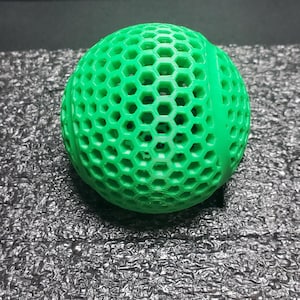 May include: A bright green, spherical object with a honeycomb pattern. The ball is made of a 3D-printed material and sits on a textured, dark surface. The design features numerous hexagonal openings throughout its surface.