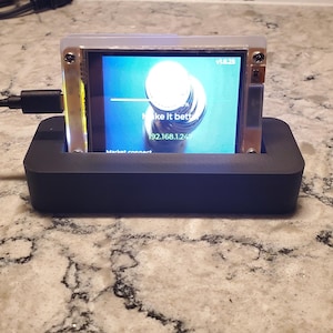 May include: A small electronic device with a screen displaying the text "Make it better" and an IP address. The device is in a black base and has a silver frame. The background is a light-colored countertop.