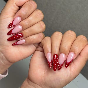 Pode incluir: Duas mãos exibindo unhas longas em formato de amêndoa. As unhas são pintadas de vermelho vibrante com bolinhas brancas. Algumas unhas apresentam uma base rosa com ponta vermelha com bolinhas. Uma pulseira dourada é visível em um pulso.