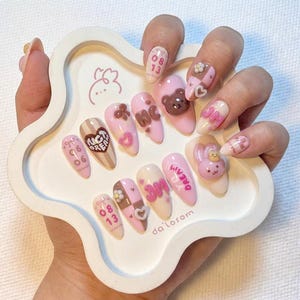 May include: A collection of almond-shaped false nails in pink, white, and brown. The nails are decorated with hearts, bows, and a teddy bear. Some nails have text, including "DREAM" and "da'losom".