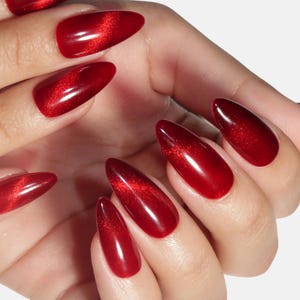 Peut inclure: Une main présentant une manucure avec des ongles artificiels rouges en forme d'amande. Les ongles ont une finition brillante avec un effet chatoyant œil de chat. La couleur est un rouge profond et riche.