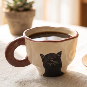 May include: A cream ceramic mug with a black cat design, filled with dark coffee. The mug has a brown handle and rim. A croissant sits on a small plate beside the mug. A succulent plant is in the background.