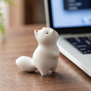 May include: A small, white ceramic squirrel figurine with a curved tail, standing upright. The squirrel has black eyes and pink accents on its ears and nose. It is positioned on a wooden surface near a laptop.
