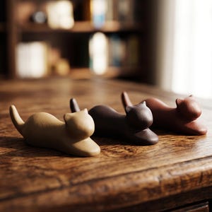 May include: Three ceramic cat figurines in tan, dark brown, and reddish-brown hues are arranged on a wooden surface. The figurines are in a resting pose, with their tails curved. The background includes a bookshelf and a window.