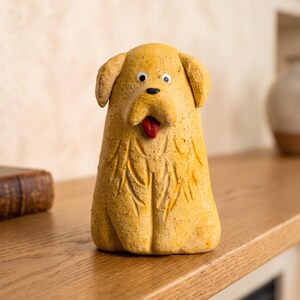 Könnte beinhalten: Gelbe Keramik-Hundefigur mit roter Zunge. Der Hund hat schwarze Augen und ein strukturiertes, geschnitztes Design auf der Brust. Die Figur steht auf einer Holzoberfläche.