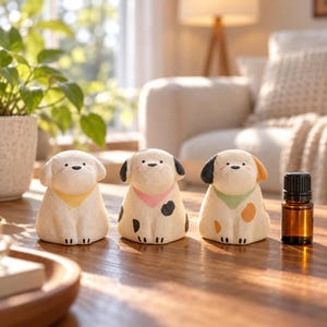 May include: Three small, cream-colored ceramic dog figurines sit in a row. The first has a yellow bandana, the second has a pink bandana and black spots, and the third has a green bandana and orange spots. A small amber glass bottle stands nearby.
