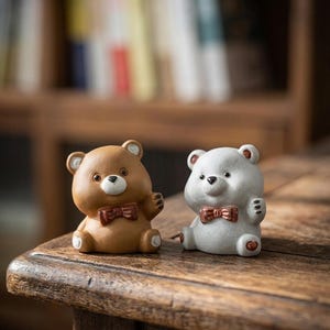 May include: Two ceramic teddy bear figurines. One is brown, the other is white. Both bears have a red bow tie and are sitting with their arms raised. The bears are on a wooden surface.