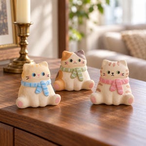 May include: Three small cat figurines made of a textured, matte material. The left cat is cream-colored with a blue scarf, the center cat is calico with a green scarf, and the right cat is tan with brown stripes and a pink scarf.