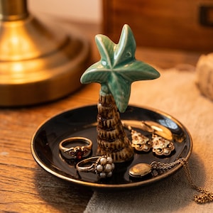 Op de afbeelding: Een zwart keramisch sieradenbakje met een gouden rand en een palmboomaccent. Het bakje bevat ringen, oorbellen en een ketting. De palmboom is groen en bruin.