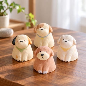 Op de afbeelding: Vier kleine, getextureerde hondjes in verschillende tinten beige, crème en bruin. Elke puppy heeft een rond lichaam, een eenvoudig geschilderd gezicht met zwarte ogen en neus, en een kleine gekleurde halsband of bandana in blauw, groen of geel.
