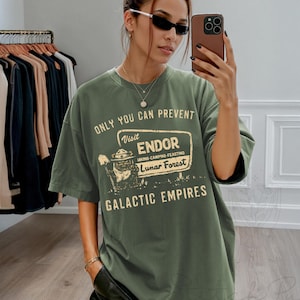 May include: An olive green t-shirt featuring the text "ONLY YOU CAN PREVENT" above a sign that reads "Visit ENDOR Hiking-Camping-Feasting Lunar Forest GALACTIC EMPIRES". The shirt is oversized with short sleeves.