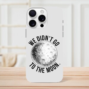 May include: White phone case with a black and white illustration of the moon. The text "WE DIDN'T GO TO THE MOON" is printed around the moon graphic. The case has a camera cutout and is on a wooden surface.