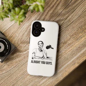 May include: White phone case featuring a black and white illustration of a woman with a microphone. The text "ALRIGHT YOU GUYS." is printed below the illustration. The phone case is on a wooden surface.
