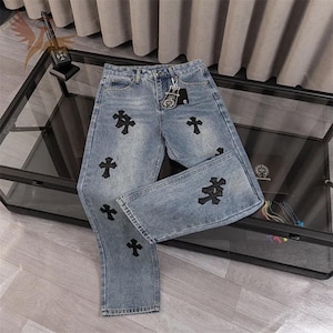 May include: Light blue denim jeans with black cross appliqués. The jeans have a straight leg cut and a classic five-pocket design. A silver keychain hangs from the belt loop. The jeans are displayed on a glass table.
