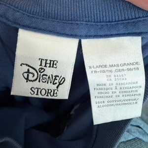 Pu&ograve; includere: Un capo blu con due etichette interne bianche. L'etichetta a sinistra mostra il logo The Disney Store in nero. L'etichetta a destra riporta la taglia X-Large, RN 84167, CA 25083, Made in Singapore e la composizione 100% cotone.