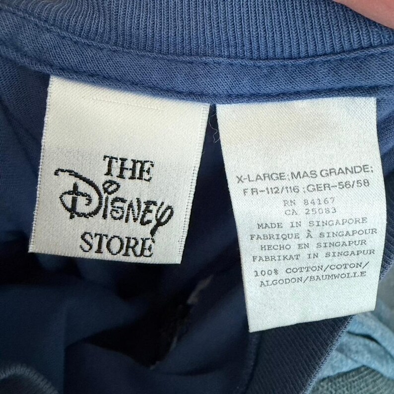 Pu&ograve; includere: Un capo blu con due etichette interne bianche. L'etichetta a sinistra mostra il logo The Disney Store in nero. L'etichetta a destra riporta la taglia X-Large, RN 84167, CA 25083, Made in Singapore e la composizione 100% cotone.