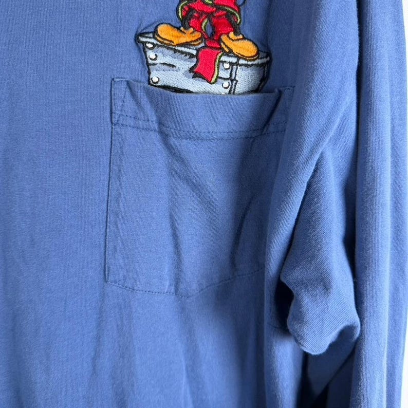 Pu&ograve; includere: Una maglia a maniche lunghe color blu pervinca con taschino sul petto. Sopra la tasca &egrave; presente un ricamo raffigurante un personaggio dei cartoni animati con scarpe gialle e pantaloni rossi, in piedi su una piattaforma grigia con rivetti.