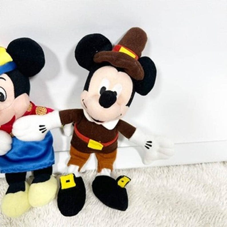 May include: Plush Mickey Mouse doll wearing a brown pilgrim outfit with a matching hat. The doll has black ears, a white face, and black shoes with yellow buckles. The doll is standing on a white surface.