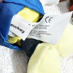 May include: Close-up of a plush toy with a blue hat, yellow and black accents, and a white tag. The tag reads "Mickey" and includes the CE mark and other text in multiple languages. The toy is made in China.