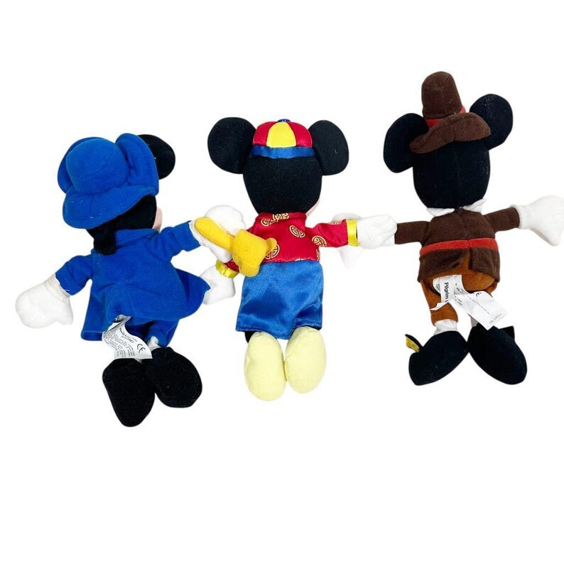 May include: Three Mickey Mouse plush toys in various outfits. One wears a blue outfit and hat, another a red and blue outfit with a yellow hat, and the third a brown outfit and hat. All have black ears, white gloves, and black shoes.