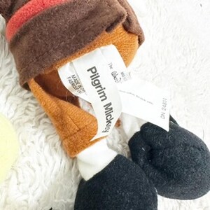 May include: A plush toy of Pilgrim Mickey Mouse, featuring a brown hat with red trim, a tan face, and black feet. The toy has a white tag with the text "Pilgrim Mickey".